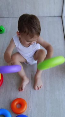 Beyaz tulumlu şirin çocuk panoramik pencerenin yanında yerde oturuyor. Çocuk oyuncak piramidin yüzükleriyle oynuyor ve onları yere vuruyor. Üst Manzara. Dikey video