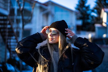 Kış güneşinde güneşlenen hoş bir kız. Kışın sıcak bir ceket, şapka ve güneş gözlüğü takan genç bir kadın dışarıda güneşli havanın tadını çıkarıyor..