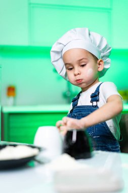 Genç aşçı parlak bir mutfakta hazırlanıyor. Şef şapkalı bir çocuk mutfak tezgahında malzemelerini karıştırıyor, yaratıcılık ve yemek pişirme hevesi sergiliyor..