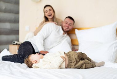 Yatakta sıcak aile zamanı. Bir çocuk yatakta huzur içinde yatarken ebeveynler gülümseyip birlikte güzel vakit geçiriyorlar..