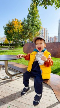 Mutlu çocuk güneşli parkta atıştırmanın tadını çıkarıyor. Neşeli bir çocuk canlı bir parkta bankta oturuyor, renkli bir ceket giyiyor ve mavi gökyüzünün altında ziyafet çekiyor..