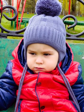 Yedekteki çocuk, ciddi bir bakış. Gri ponpon şapkalı genç bir çocuk bankta oturuyor, açık havada parlak kırmızı ve mavi bir ceketle ciddi görünüyor..