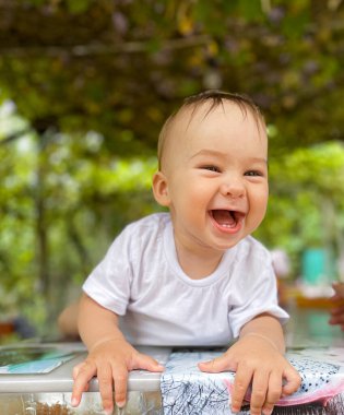 Gülen bebek dışarıda eğleniyor. Neşeli bir bebek dışarıda yeşil bir gölgeliğin altında oynarken gülüyor, güneşli atmosferin ve sıcak günün tadını çıkarıyor..