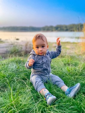 Göl kenarında doğanın tadını çıkaran bir bebek. Küçük bir çocuk arka planda yumuşak bir akşam ışığı olan sakin bir gölün yanındaki yeşil çimlerde oturur..