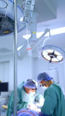 Kürsüdeki tuz solüsyonuna serum bağlanmış. Ameliyathane ekipmanı. Erkek cerrahlar arka planda ameliyat yaparlar. Dikey video.