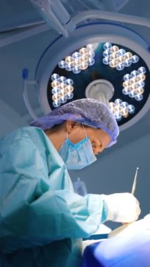 Şapka, maske ve bornoz takan ve elinde metal bir alet tutan kadın doktor. Ameliyatta çalışan cerrahın görüş açısı düşük. Dikey video.