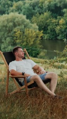 Beyaz tişörtlü beyaz sakallı beyaz adam katlanır bir sandalyede oturur. Mutlu, rahat adam gülümseyerek doğanın tadını çıkarıyor. Dikey video