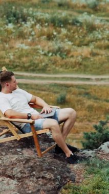 Dalgın, rahat erkek kayanın üzerindeki katlanır sandalyede oturuyor. Adam işten sonra dizüstü bilgisayarla manzaranın tadını çıkarıyor. Yüksek açı. Dikey video