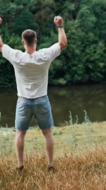 Beyaz tişörtlü ve kot şortlu güçlü kaslı bir adamın arka görünüşü. İnsan, hayatı ve doğada boş zamanı zevkle el kaldırır. Dikey video