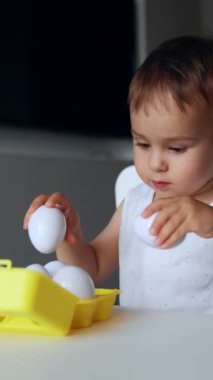 Küçük çocuk içeride, masada oturup oyun oynuyor. Sevimli çocuk beyaz plastik yumurtalarla oynuyor. Dikey video