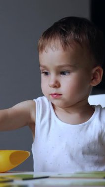 Beyaz tişört giyen bebek oyuna odaklanmış. Ciddi çocuk masasında oturmuş oyuncaklarını oynatıyor. Dikey video