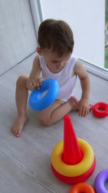Panoramik pencerenin önünde yerde oturan küçük sevimli bir oğlan. Oyuncak piramidi birleştirmeye odaklanmış bebek. Üst Manzara. Dikey video