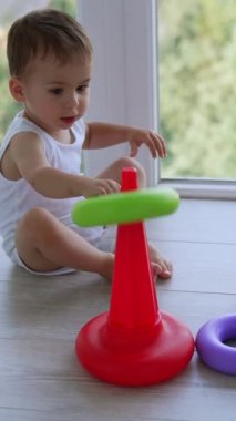 Oyuncak piramidi montajı çok güzel. Beyaz çocuk pencerenin yanında evde oynuyor. Dikey video