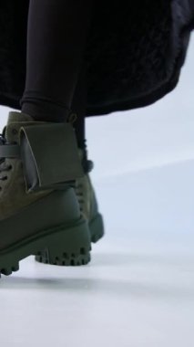 Bileklerinde haki çizme olan bir kadın. Siyah elbiseli ve uzun paltolu modelin onu stüdyoda gösteren şık ayakkabıları var. Dikey video