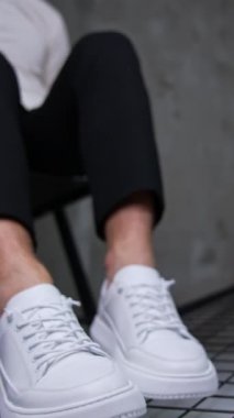 Sandalyede oturan erkek modern spor ayakkabılarını gösteriyor. Beyaz gömlekli ve siyah pantolonlu tanınmayan adam kameranın önünde beyaz spor ayakkabıları sunar. Kapatın. Arkaplan bulanık. Dikey video