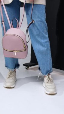 Sıradan, modern bir yaşam tarzı. Deri bir sırt çantası tutan kot ve beyaz spor ayakkabılı bir kız. Arka planda siyah perde. Dikey video