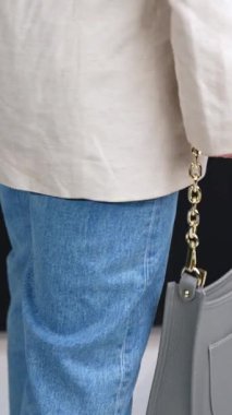 Altın zincir saplı, güzel, gri deri bir çanta. Ceketli ve kot pantolonlu bir kadın elinde modern bir çanta tutuyor. Dikey video