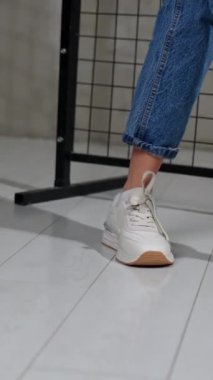 Kız arkasını döner ve rahat beyaz ayakkabıları gösterir. Kot pantolonlu bir kadın şık ayakkabılar giyer. Arka planda siyah ızgara var. Dikey video