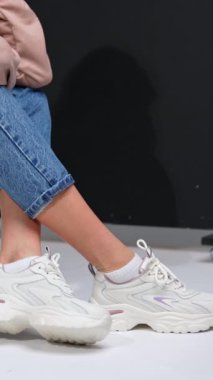 Siyah basamakta oturan bir kızın kırpılmış görüntüsü. Stüdyoda kot pantolon, sweatshirt ve beyaz spor ayakkabı mankeni. Dikey video