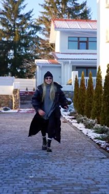 Sıcak kıyafetler giyen neşeli, uzun saçlı beyaz kadın kameraya doğru koşuyor ve neşeyle zıplıyor. Kışın açık havada enerjik bir bayan. Dikey video.