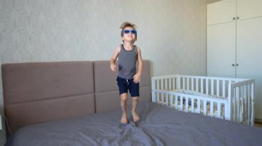 Güneş gözlüğü takan beyaz çocuk çift kişilik yatakta zıplıyor. Mutlu çocuk evde eğleniyor..