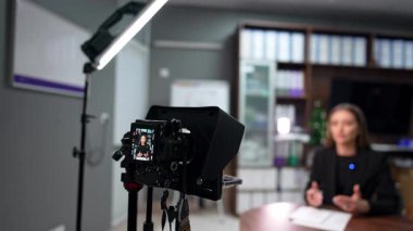 Siyah ceketli beyaz tenli çekici esmer ofiste bir blog kaydediyor. Tripod ekranındaki kamera kadın blogcuyu gösteriyor. Bulanık arkaplan.