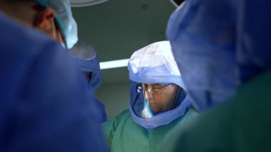 Gözlük takan, koruyucu kask takan ve cerrahi ekipte çalışan beyaz bir erkek cerrah. Doktor, travma operasyonunda meslektaşlarıyla birlikte çalışıyor..