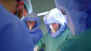 Koruyucu miğferlerle donatılmış profesyonel erkek doktorlar bir hastayı ameliyat ediyor. Baş cerrah metal alet uyguluyor.