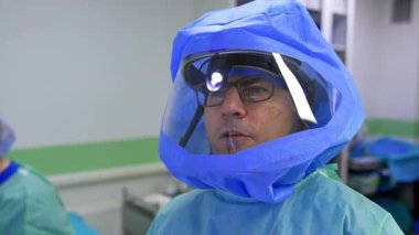 Yetişkin, beyaz, gözlüklü bir cerrah. Farlar ve koruyucu miğfer. Doktor ameliyathanede kamerayla konuşuyor. Kapat..
