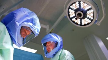 Koruyucu kasklı beyaz cerrahlar ameliyata gülümsüyor. Yuvarlak lambanın altında çalışan doktorlara düşük açılı bakış açısı. Düşük açı görünümü.