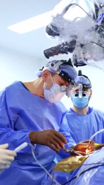 Cerrahi hoparlörlerdeki özenli erkek cerrah kan emme aletini tutuyor ve enstrümanı bekleyen asistana elini uzatıyor. Mikroskop ameliyat doktorunun üstünde. Dikey video.