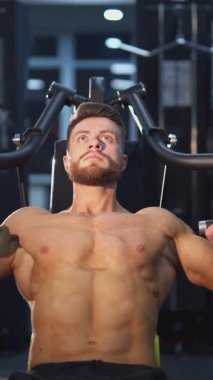 Simülatördeki sporcu. Modern spor merkezinde kaslı vücut egzersizi yapan tişörtsüz yakışıklı bir adam. Beden eğitiminde konsantre vücut geliştirme portresi. Dikey video