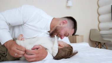 Sakallı adam sarılıyor, öpüyor ve gıdıklıyor. Yatakta tatlı bir oğlan çocuğu var. Sevgi dolu baba neşeli oğluyla oynuyor..
