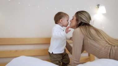 Beyaz tişörtlü ve haki pantolonlu küçük bebek annesine doğru geliyor. Neşeli çocuk yatağın yumuşak yastıklarına atlar..