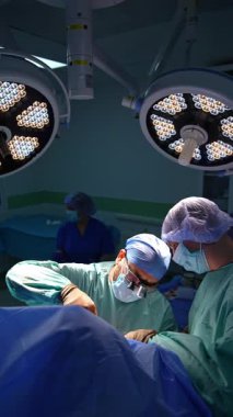 Erkek cerrahlar iki yuvarlak lambanın altında ameliyat yaparlar. Sakallı doktor iş yerinde metal aletler kullanıyor. Dikey video.