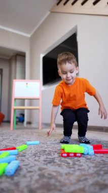 Beyaz çocuk turuncu tişört ve siyah pantolon giyerek evde oynuyor. Blokları olan çocuk binasını düşük açılı görüyor. Kara kedi arka planda yürüyor. Dikey video.