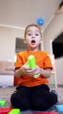 Turuncu tişört ve siyah pantolon giyen beyaz erkek bebek yerde oturuyor. Mutlu çocuk renkli tuğlalarla oynuyor. Düşük açı. Dikey video.