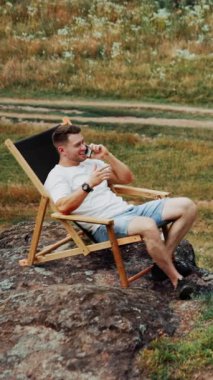 Kayanın üzerinde katlanan sandalyede oturan orta yaşlı bir adam. Erkek girişimci telefonda konuşur, doğayla uzaktan yakından ilgilenir. Dikey video