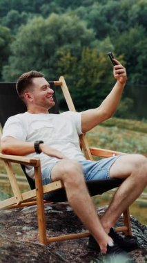 Neşeli, gülümseyen, beyaz bir adam telefonuyla sohbet ediyor. İnsan doğada katlanır sandalyede oturur. Dikey video