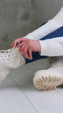 Beyaz kazak giyen ve kot giyen kadın model yerde oturuyor. Kadın traktör tabanında şık beyaz çizmelerine dokunuyor. Kapatın. Dikey video