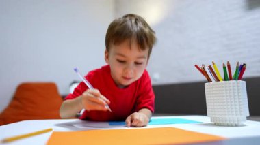 Kırmızı tişörtlü neşeli çocuk elinde bir kalem tutuyor. Güzel bebek evde neşeyle çiziyor. Düşük açı görünümü.