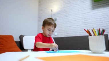 Masada oturan geveze, beyaz bir çocuk kalemle resim çiziyor. Neşeli çocuk renkli kağıda resim çizer..