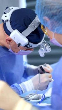 Mavi şapkalı, farlı ve maskeli bir erkek cerrah hastaya doğru eğildi. Doktor göğüs büyütme ameliyatında elektrikli alet uyguluyor. Dikey video.