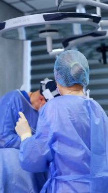 Sarı gözlüklü hemşire doktor için metal bir alet tutuyor. Erkek cerrah göğüs büyütme ameliyatı yapıyor. Dikey video.