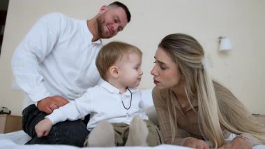 Yatakta oturan sevimli erkek bebek annesine bakıyor. Anne ve oğul burunlarına sevgiyle dokunur. Kapatın. Sakallı, gülümseyen adam arka planda oturur. Düşük açı görünümü.