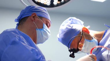 Operasyonu şapkalı ve maskeli cerrahlar gerçekleştirir. Asistanın elinde kan emici bir alet var. Doktor kullanılmış bir aleti hemşireye verir. Düşük açı görünümü.