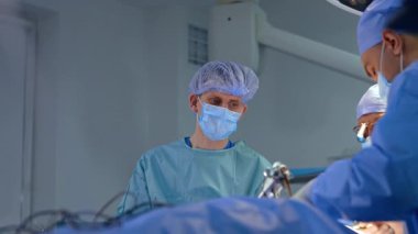 Ameliyat masasında duran erkek ameliyat hemşiresi. İki cerrah tarafından yapılan ameliyatı izleyen doktor. Düşük açı görünümü.