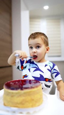 Sevimli beyaz çocuk bütün pastayı çatalla yiyor. Mutlu çocuk mutfakta tatlıyı seviyor. Dikey video.