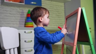 Mavi kazak giyen tatlı çocuk tahtaya kırmızı kalemle çiziyor. Evde sanat hobisi olan aktif bir çocuk. Yan görünüm.