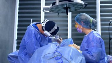 Mavi bornozlu doktor metal aletlerle hastanın üzerine eğildi. Sarı koruyucu gözlük takan ameliyat hemşiresi göğüs ameliyatında doktora yardım ediyor..